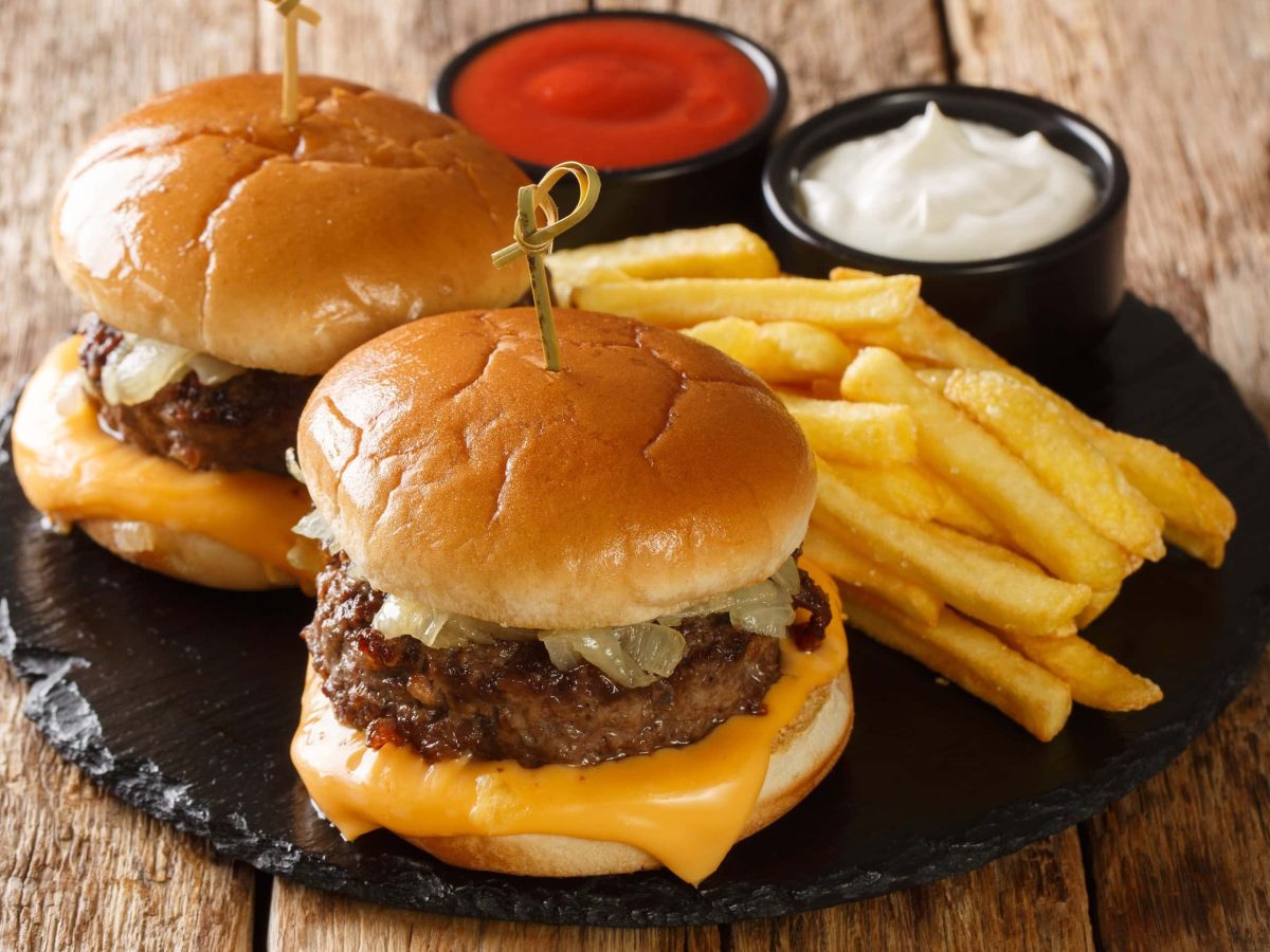 Zwei Wisconsin Butter Burger mit Pommes und je einer Schale Mayo und Ketchup auf Schieferplatte auf Holz. Draufsicht.