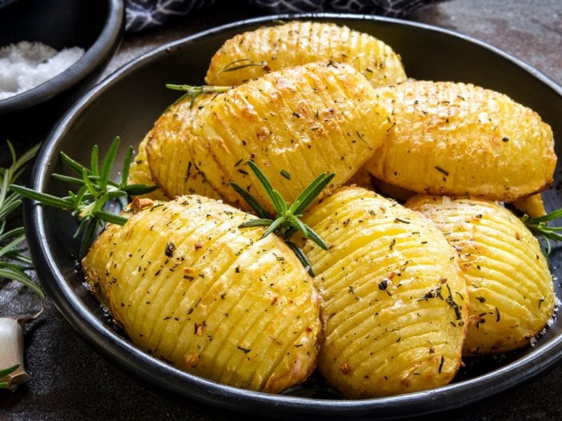 Ein Teller zitronige Hasselback-Kartoffeln mit Rosmarin, daneben Rosmarinzweige und Knoblauchzehen.