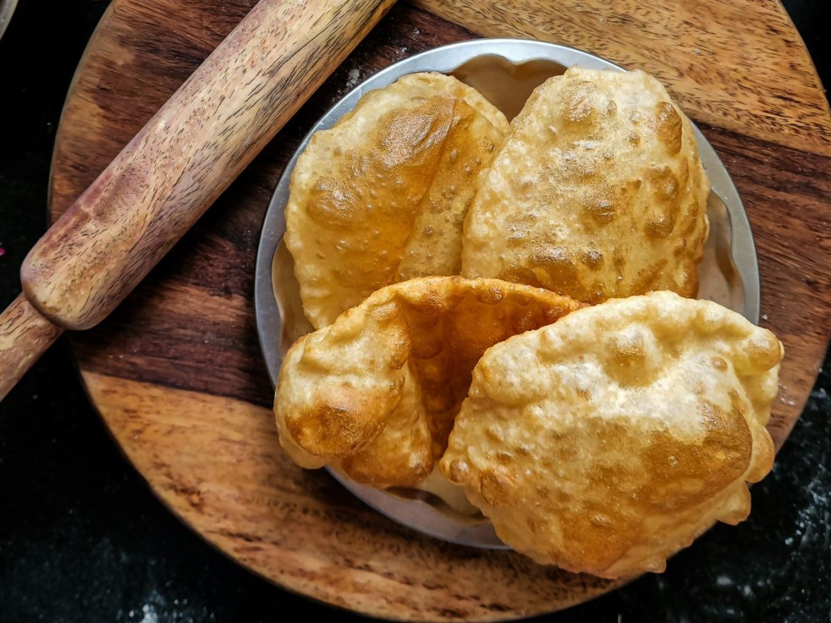 Bhatura selber machen: Mehrere Bhatura in einer Schüssel neben einem Nudelholz auf einem Holzbrett vor dunklem Hintergrund.