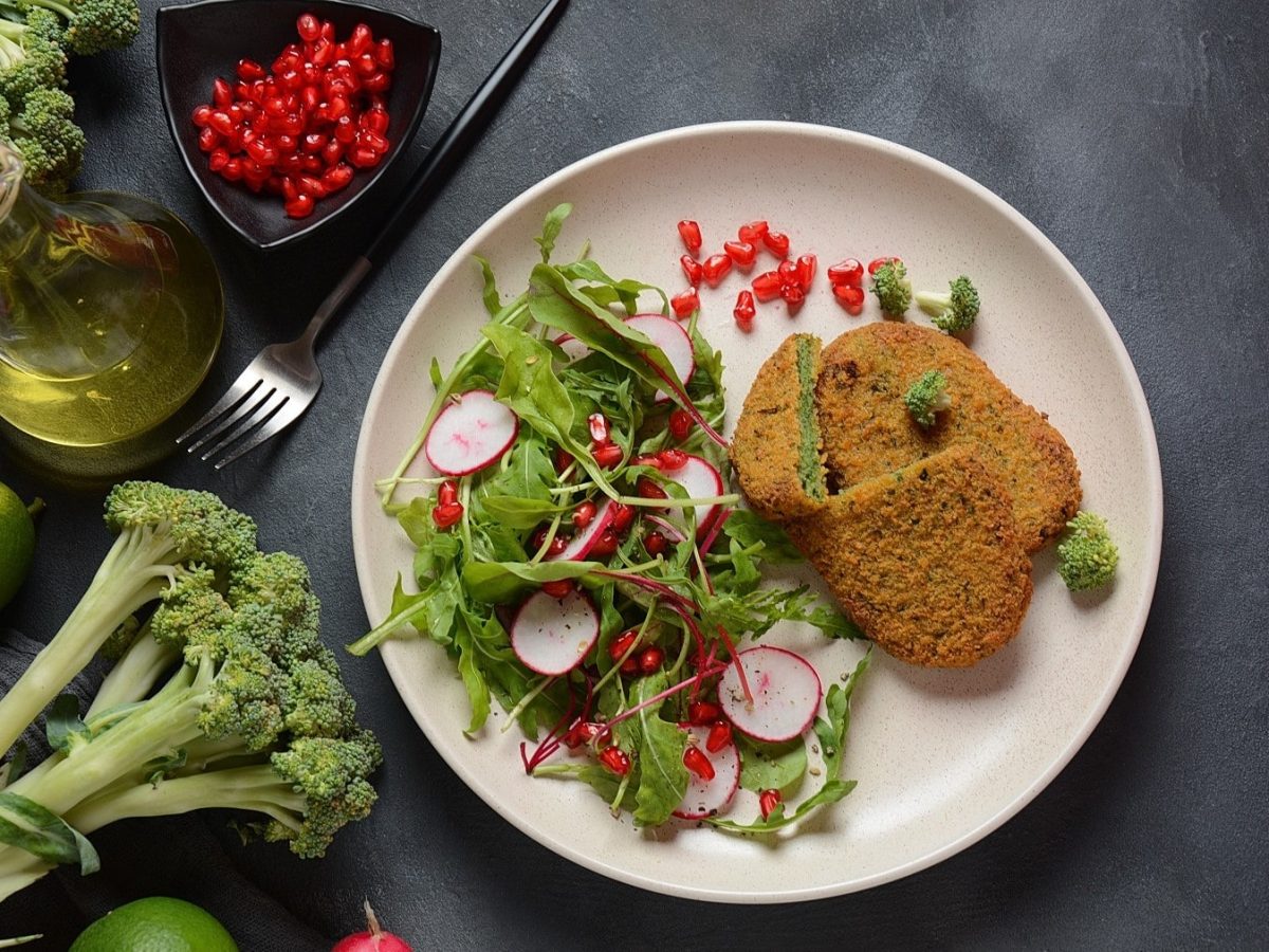 Brokkoli-Schnitzel mit Rucola-Radieschen-Salat, garniert mit frischer Vinaigrette und Granatapfelkernen, serviert auf einem weißen, flachen Teller auf einem steinernen Tisch.