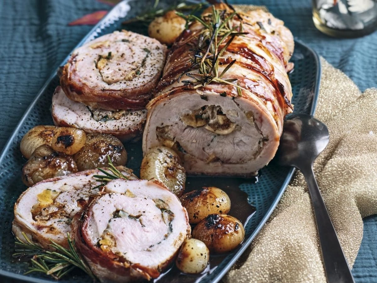 Gerollter Truthahnbraten gefüllt mit Feigen und Kräutern. Umwickelt mit Speck und garniert mit Rosmarin. Auf einem blauen Teller mit karamellisierten Zwiebeln serviert.