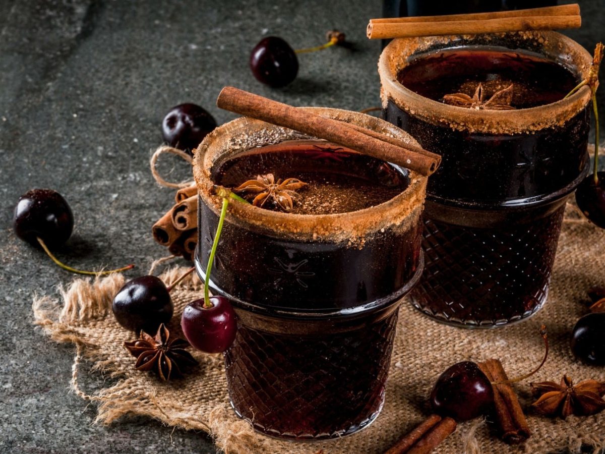 Zwei Gläser Glühwein mit Amaretto und Kirschen mit Zimtrand dekoriert mit Zimtstangen und Kirschen vor dunklem Hintergrund.