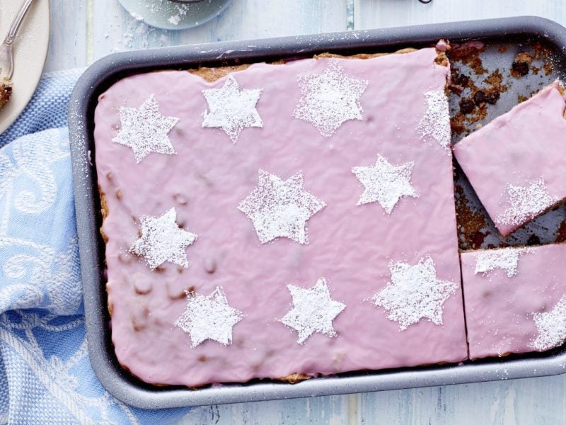 Glühweinkuchen vom Blech in der Form mit rosa Zuckerguss und Puderzuckersternen verziert vor blauem Hintergrund.