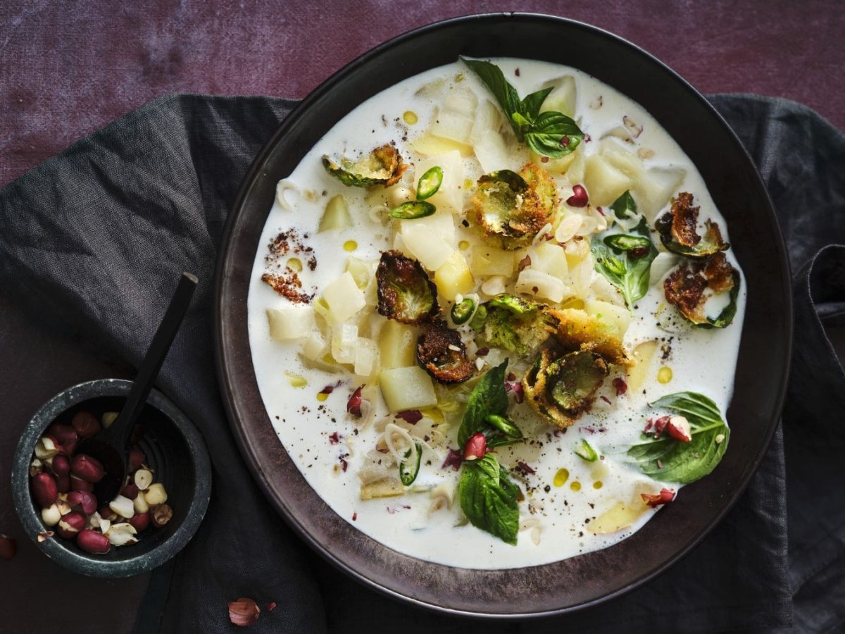 Kohlrabi-Kokos-Suppe mit paniertem Rosenkohl, Erdnüssen, Chiliflocken und frischem Thaibasilikum, serviert in einer dunklen Keramikschale auf dunklem Untergrund.