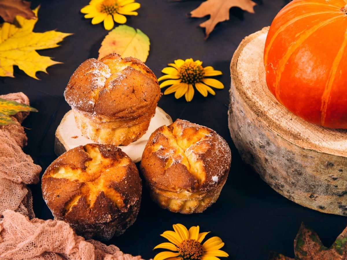 Drei herzhafte Kürbismuffins vor dunklem Hintergrund mit Herbstblättern, einem Kürbis und gelben Blüten als Deko.