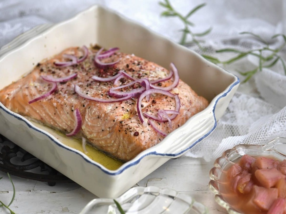 Lachs in einer eckigen weißen Auflaufform. Garniert mit roten Zwiebeln und Pfeffer. Rechts daneben befindet sich eine kleine Glasschale mit Rhabarberkompott. Frische Estragonzweige sind dekorativ um die Speisen drapiert.