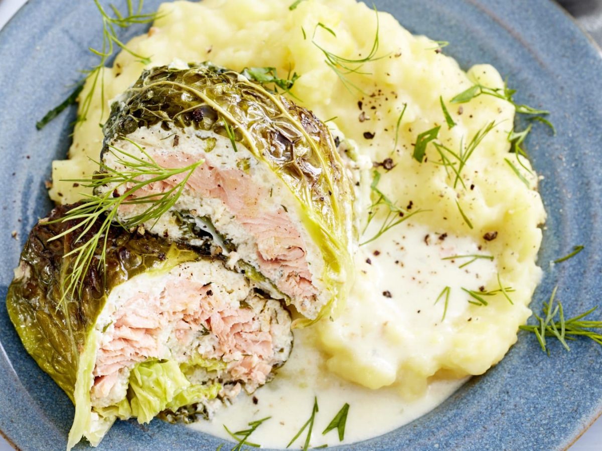 Lachs-Wirsing-Wickel mit Kartoffelbrei und Dill auf einem blauen Teller vor hellem Hintergrund.