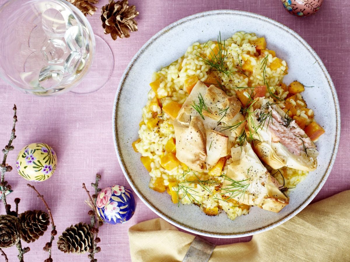 Lachs-Päckchen auf Kürbis-Risotto, garniert mit frischem Dill und serviert auf einem weißen Teller auf rosanem Untergrund.