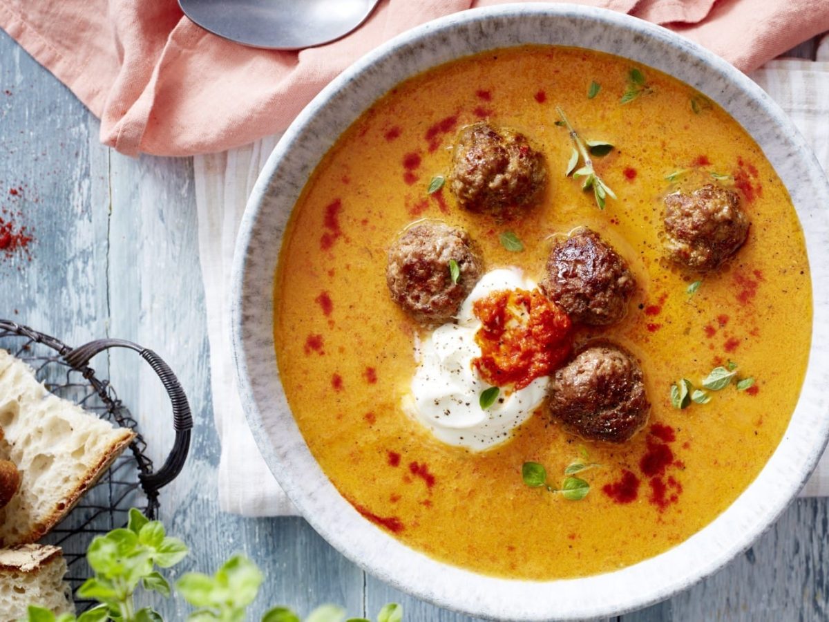 Paprikasuppe mit Hackbällchen, garniert mit Schmand, Ajvar und frischem Oregano und serviert in einer Suppenschüssel auf einem rustikalen Holztisch.