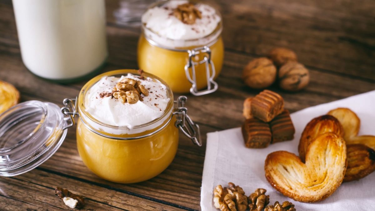 Zwei Einmachgläser mit Kürbis-Walnuss-Shake mit Sahne, daneben verschiedene Kekse vor Holzhintergrund.