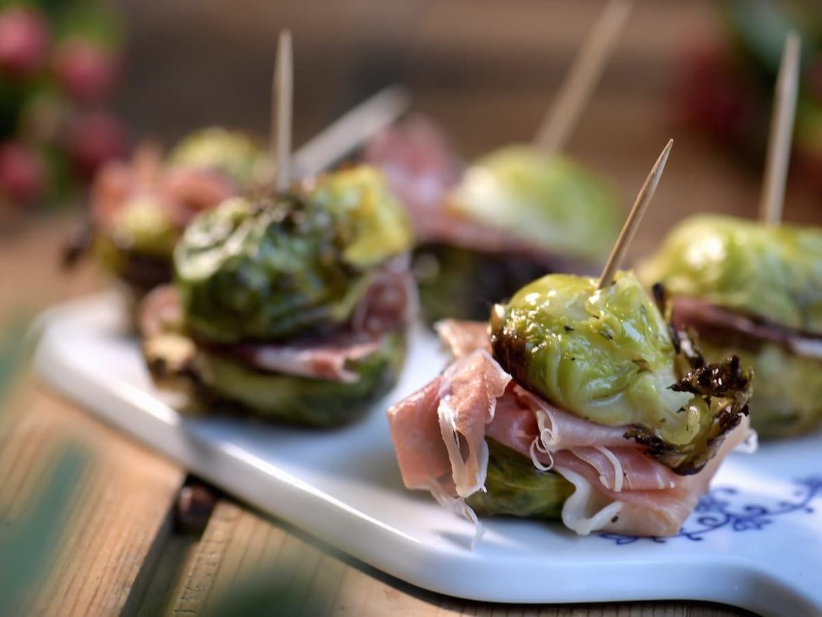 Halbierter Rosenkohl mit Salami und Parmaschinken, fixiert mit einem Zahnstocher auf einem weißen Brett serviert