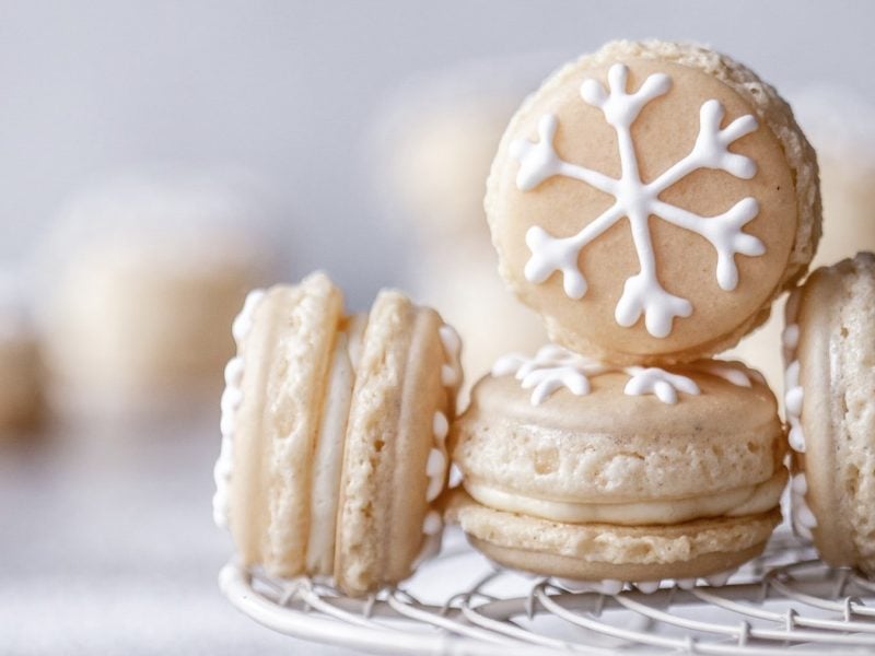 Einige Schneeflocken-Macarons auf einem Gitter platziert vor hellem Hintergrund mit weiteren Macarons verschwommen im Hintergrund.