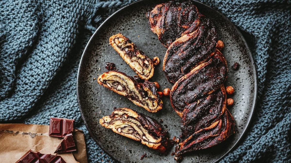 Draufsicht: Geflochtene Schokoladen-Nuss-Babka auf einem grauen Steingutteller. Links daneben Schokoladenstücke.