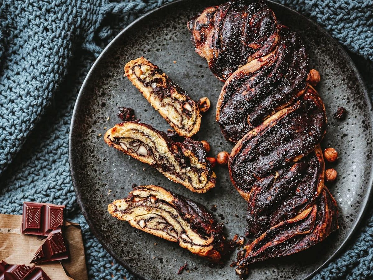 Draufsicht: Geflochtene Schokoladen-Nuss-Babka auf einem grauen Steingutteller. Links daneben Schokoladenstücke.