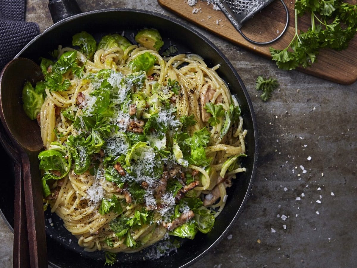 In einer Pfanne sind Spaghetti mit Rosenkohl, Speck, Parmesan und Petersilie angerichtet. Sie sind mit Käse bestreut.