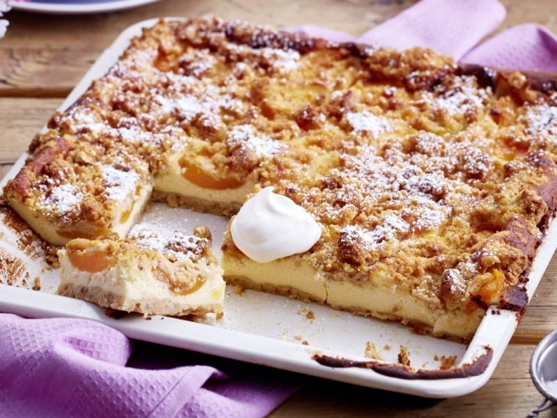 Aprikosen-Schmand-Kuchen im Blech auf einem lila Tuch vor Holzhintergrund.