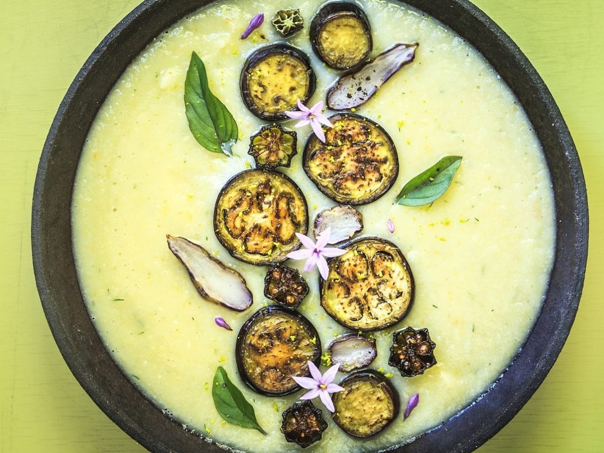 Auberginensuppe mit getrocknetem Gemüse, Thai-Basilikum und gerösteten Auberginenscheiben, in einer dunklen Schale auf gelbem Untergrund serviert.