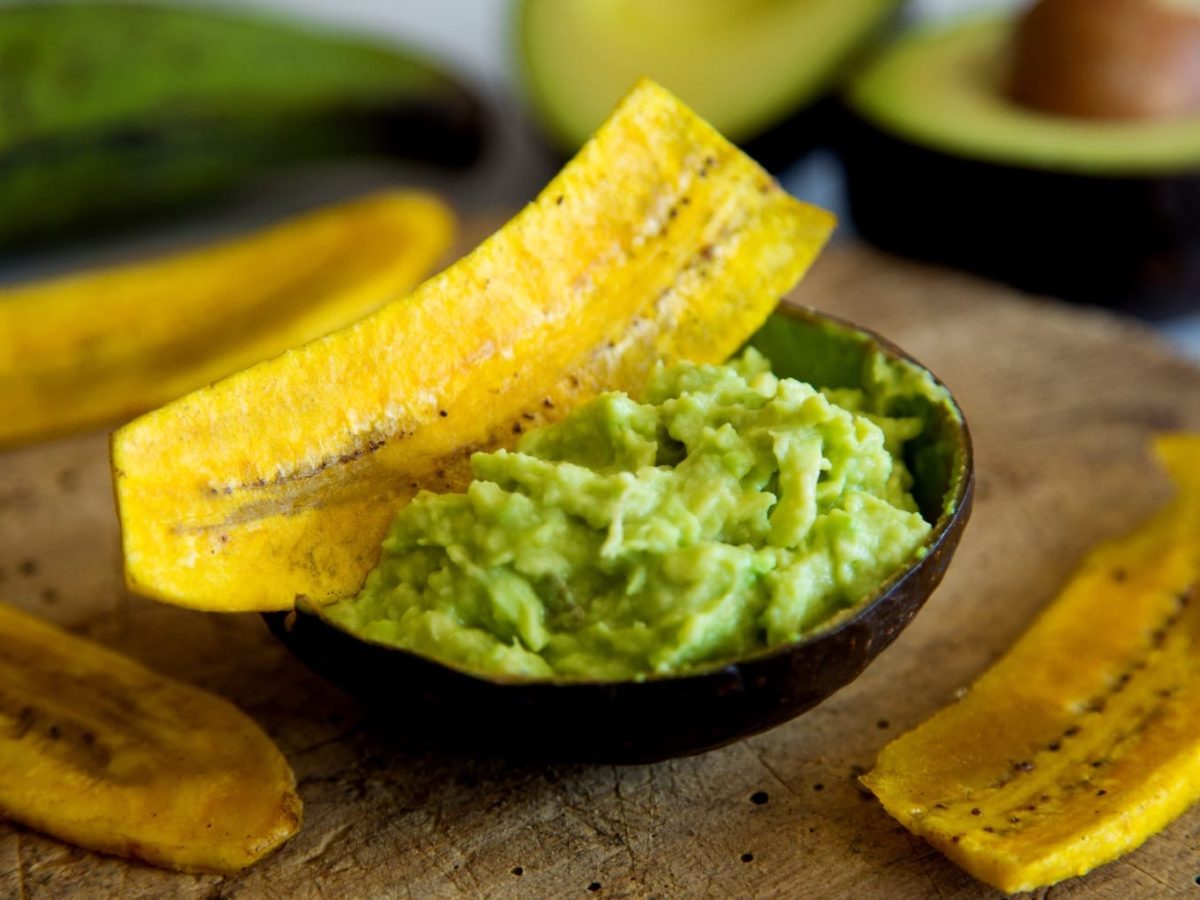 Einige Bananenchips auf einem Holzbrett mit der Mojo-Avocado in der Avocado-Schale.