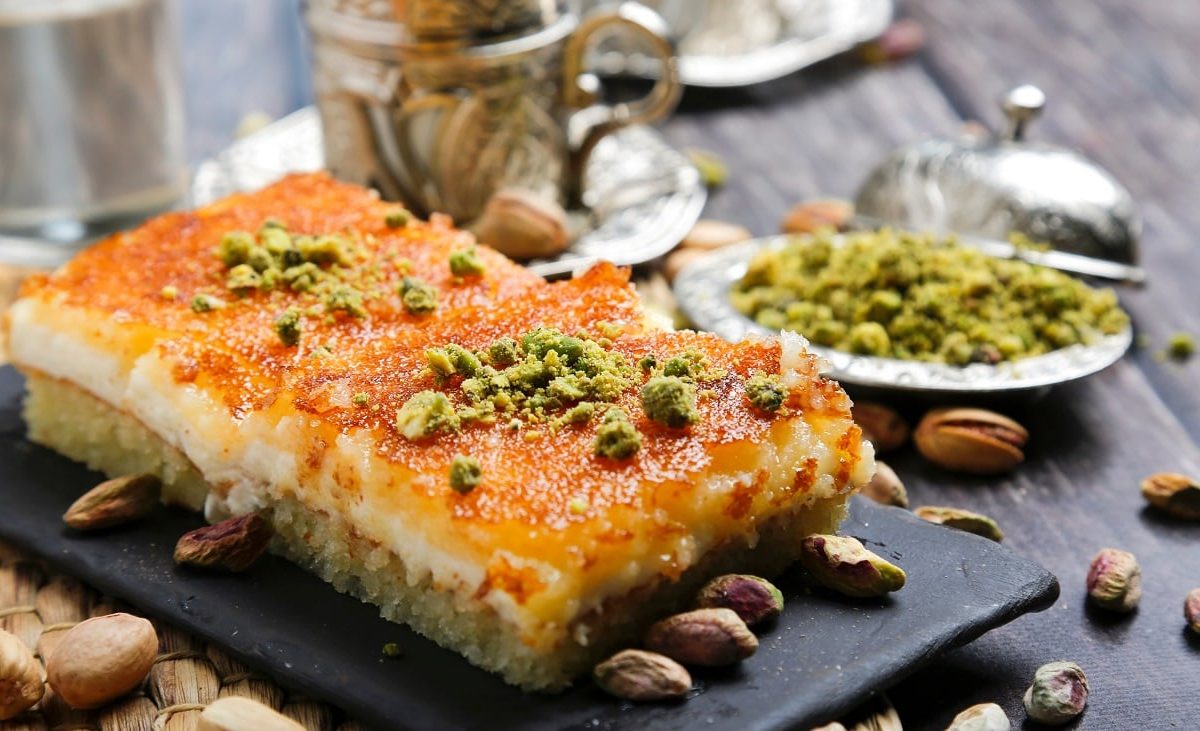 Frontalsicht: zwei flasche kunchenstücke liegen auf einem Untergrund aus Schiefer. die kuchen sind mit Pistaziem berstreut und haben eine feuchte Oberfläche. Auch frumherum liegen viele Pistazien.