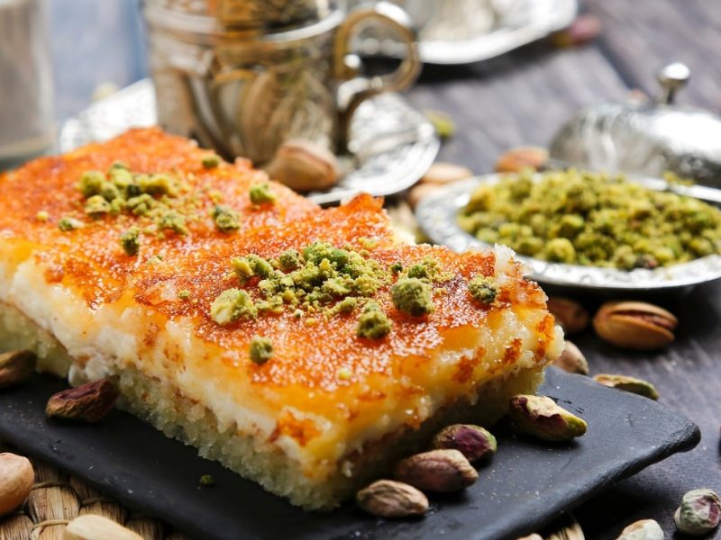 Frontalsicht: zwei flasche kunchenstücke liegen auf einem Untergrund aus Schiefer. die kuchen sind mit Pistaziem berstreut und haben eine feuchte Oberfläche. Auch frumherum liegen viele Pistazien.