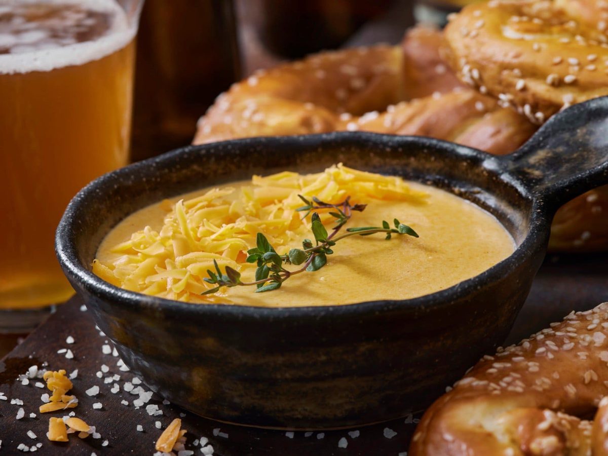 Beer Cheese Dip in einer schwarzen Schale auf Granitplatte. Im Bild außerdem Bretzeln und ein Glas Bier. Grobes Salz und Thymian im Bild verteilt. Frontalaufnahme..