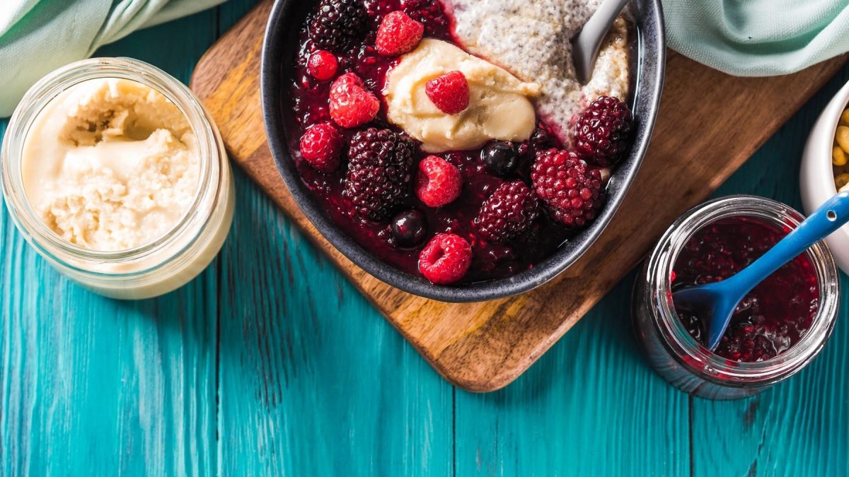 Fruchtige Rote Grütze in einer Bowl mit Chia-Pudding und Mandelmus, serviert auf einem Holzbrett auf blauem Untergrund.