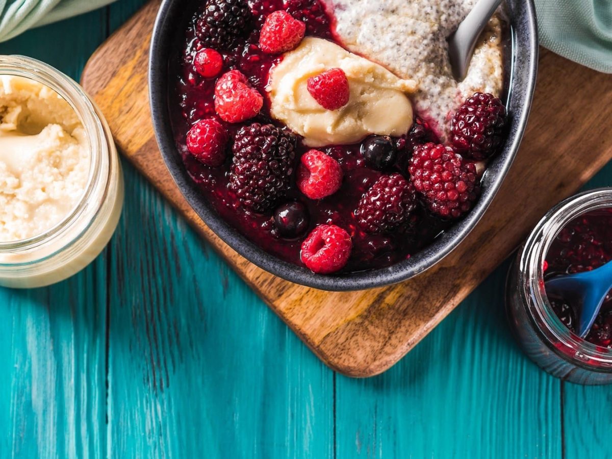 Fruchtige Rote Grütze in einer Bowl mit Chia-Pudding und Mandelmus, serviert auf einem Holzbrett auf blauem Untergrund.