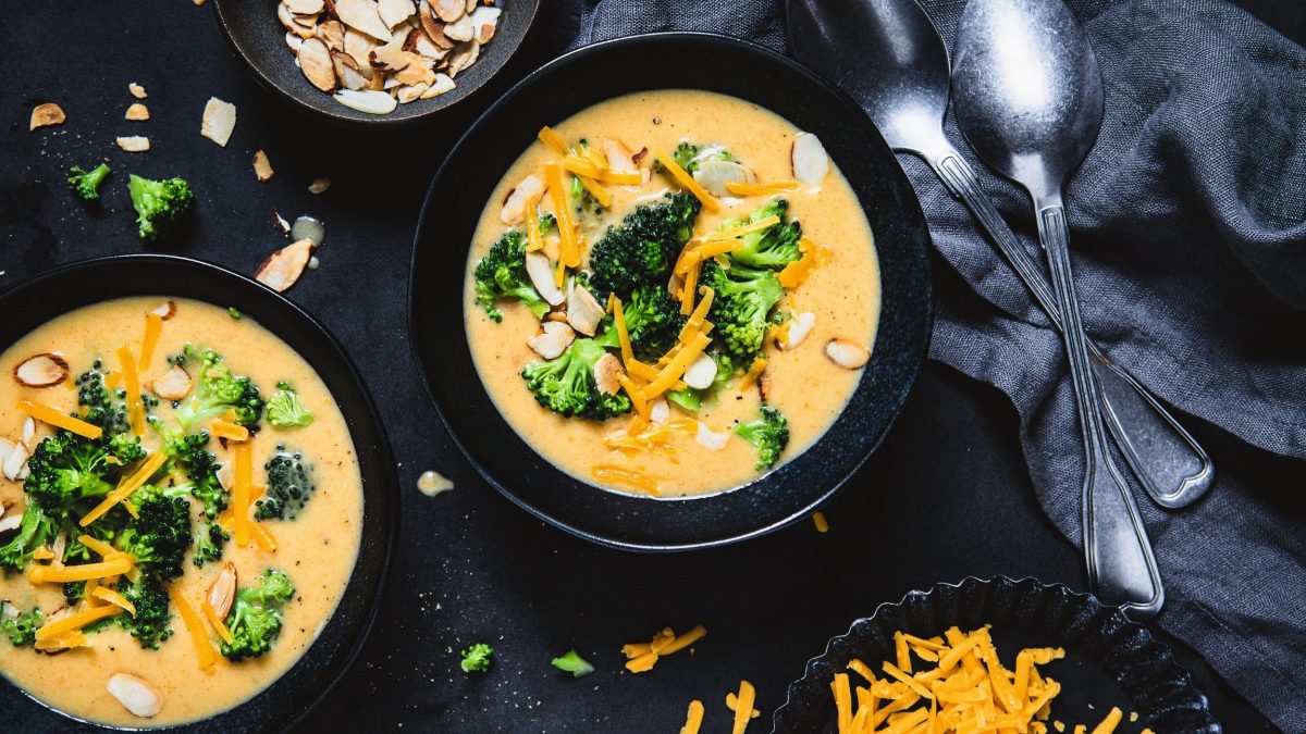 Zwei Schalen Brokkoli-Cheddar-Suppe mit Mandelblättchen in der Draufsicht, daneben zwei Löffel und Schalen mit Zutaten.