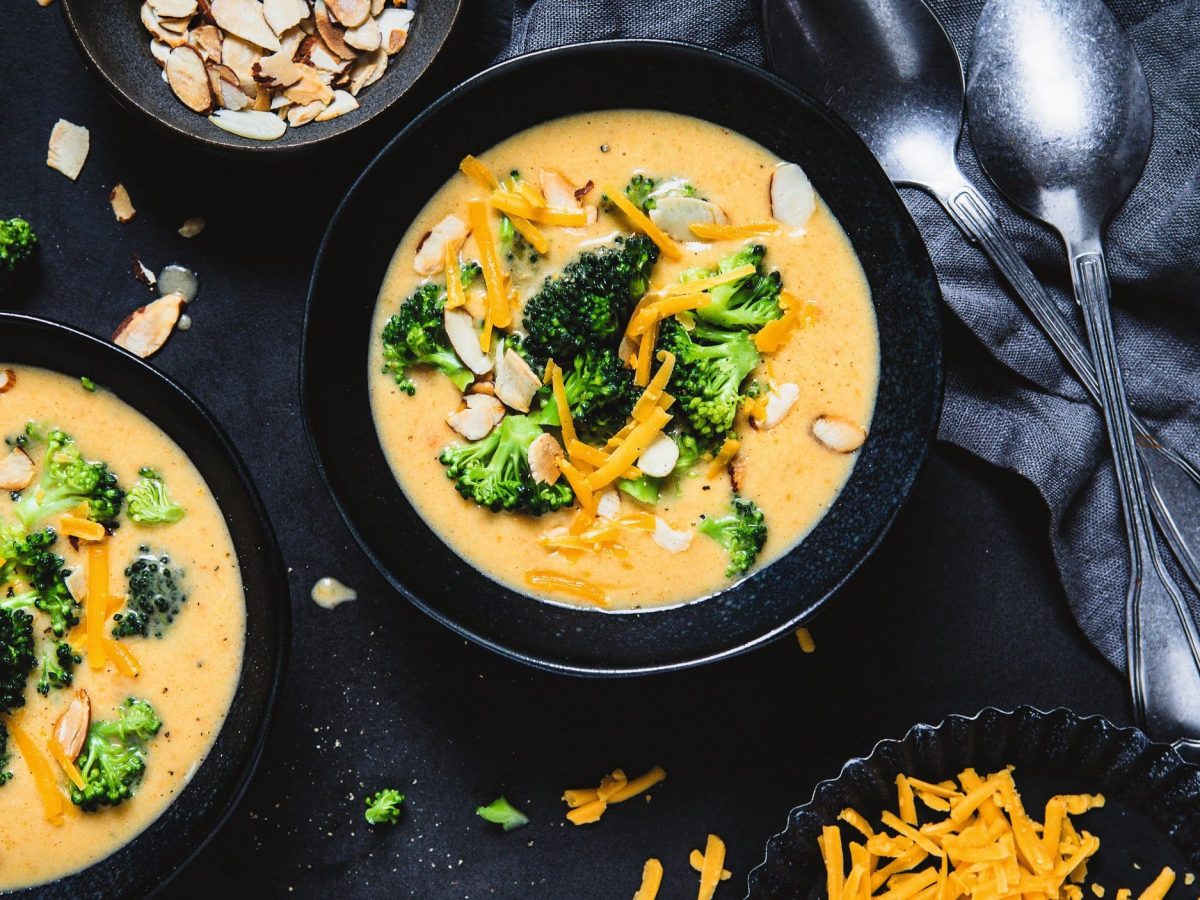 Zwei Schalen Brokkoli-Cheddar-Suppe mit Mandelblättchen in der Draufsicht, daneben zwei Löffel und Schalen mit Zutaten.