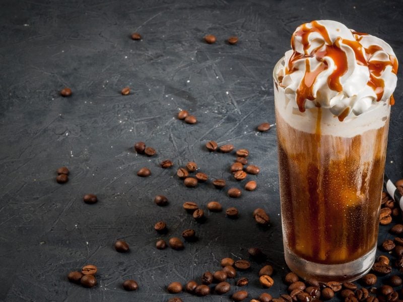 EIn Glas Caramel Macchiato mit Schlagsahne und Karamellsauce, daneben Kaffeebohnen und Strohhalme.