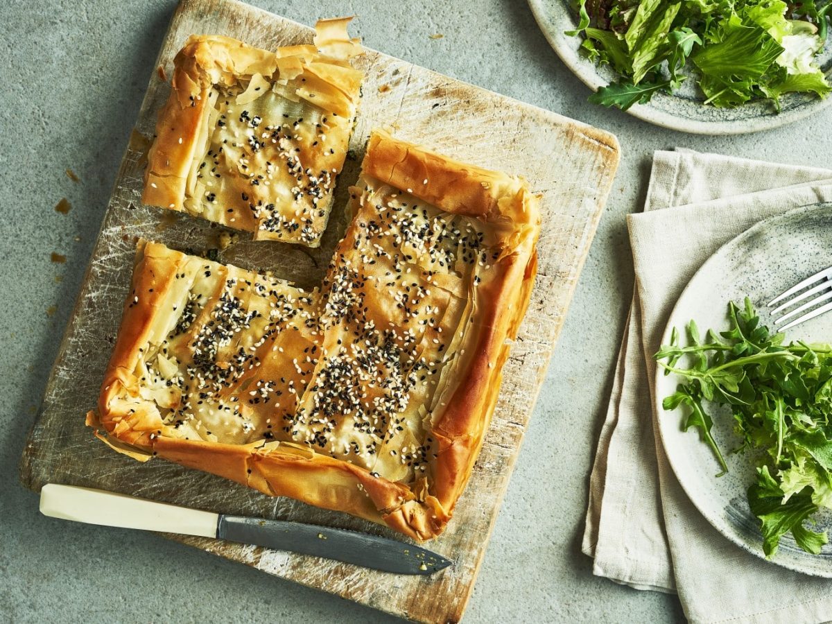 Filokuchen mit Kürbis auf einem Holzbrett in der Draufsicht, daneeben Teller mit Salat und Besteck.