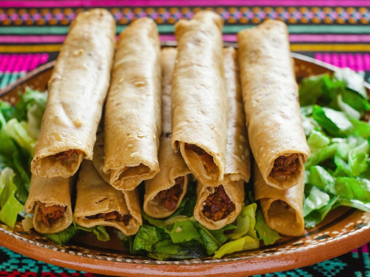 Mehrere gerollte Hühnertacos auf einem braunen Teller mit salat darunter, auf einer bunten Decke.