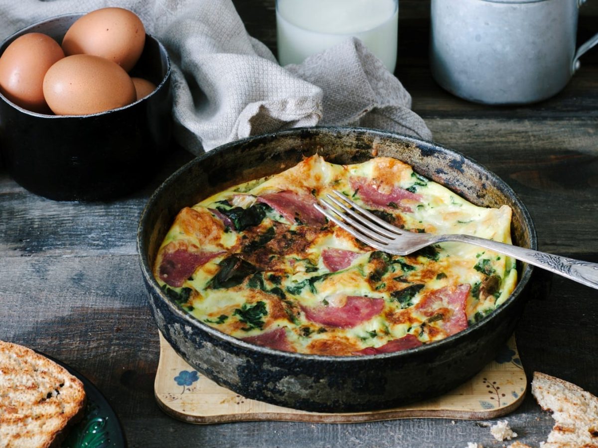 Eine Frittata mit Spinat und Schinken in einer gusseisernen Pfanne, daneben Eier und geröstetes Brot.