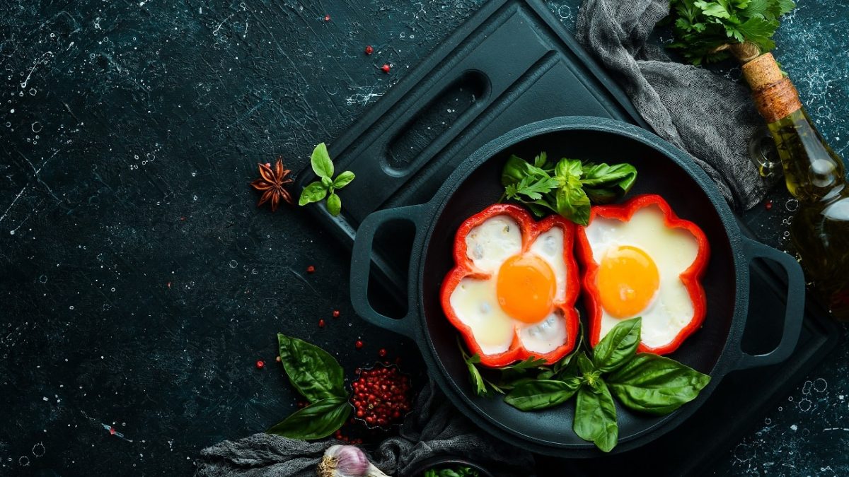 Draufsicht: zwei paprika ohne Deckel stehen in einem dunklen Topf. in den aufgeschnittenen Paprika ist heweils ein Spiegelei. Drumherum liegen Petersilie und Basilikum.