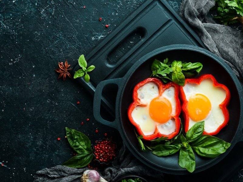 Draufsicht: zwei paprika ohne Deckel stehen in einem dunklen Topf. in den aufgeschnittenen Paprika ist heweils ein Spiegelei. Drumherum liegen Petersilie und Basilikum.