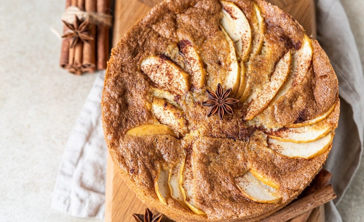 Draufsicht: ein runder Apfelkuchen, auf dem ein Sternanis liegt. Der Gewürz-Apfelkuchen liegt auf einem Bretten, daneben liegen mehrere Stangen Zimt und ein weiteres Sternanis.