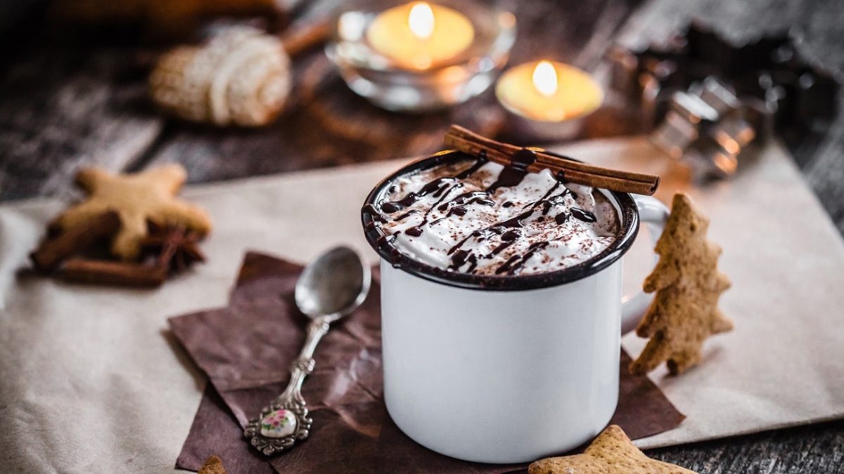 Frontalsicht: EIn weihnachtliche gedeckter Tisch mit Kerzen, Kekesen und eine Blechtasse, in der ein hesses Getränk mit einer Sahnehaube steht. Es handelt sich um eine heisse Lebkuchen-Schokolade.