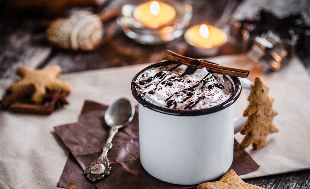 Frontalsicht: EIn weihnachtliche gedeckter Tisch mit Kerzen, Kekesen und eine Blechtasse, in der ein hesses Getränk mit einer Sahnehaube steht. Es handelt sich um eine heisse Lebkuchen-Schokolade.