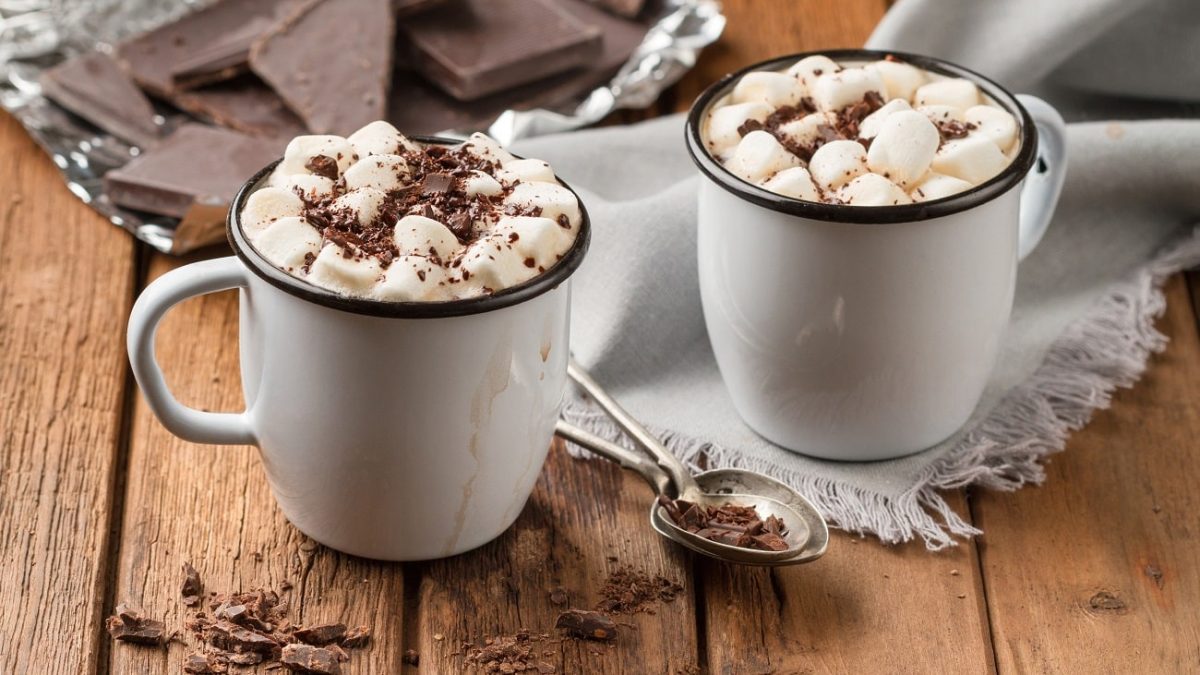 Frontalsicht: Zwei Blechtassen mit Heiße Schokolade mit Marshmallows stehen auf einem Untergrund aus Holz. Daneben liegen Schokostreusel und ein Löffel und ein Tuch.