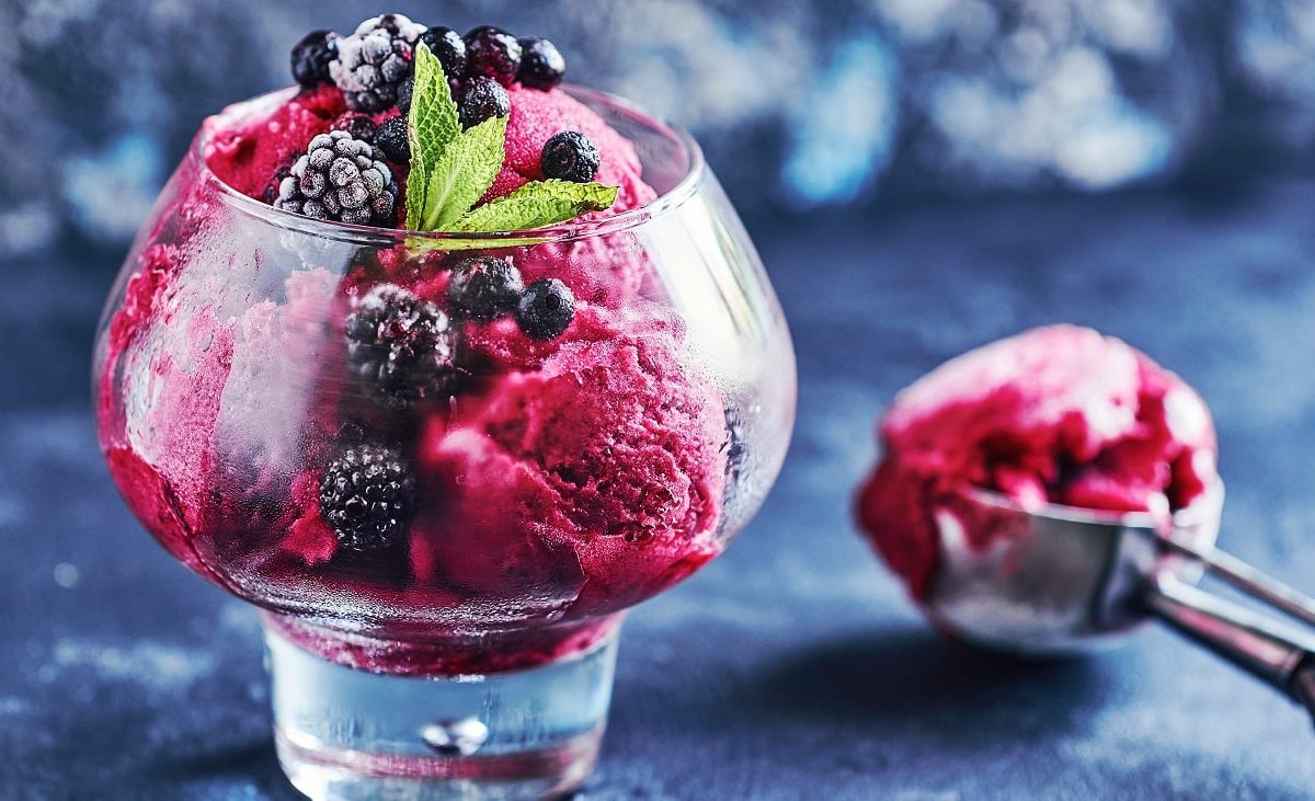 Frontalsicht: Ein Glas mit einem Johannisbeer-Brombeer-Sorbet mit Prosecco. Daneben liegt ein Eisportionierer. der Untergrund ist dunkel.