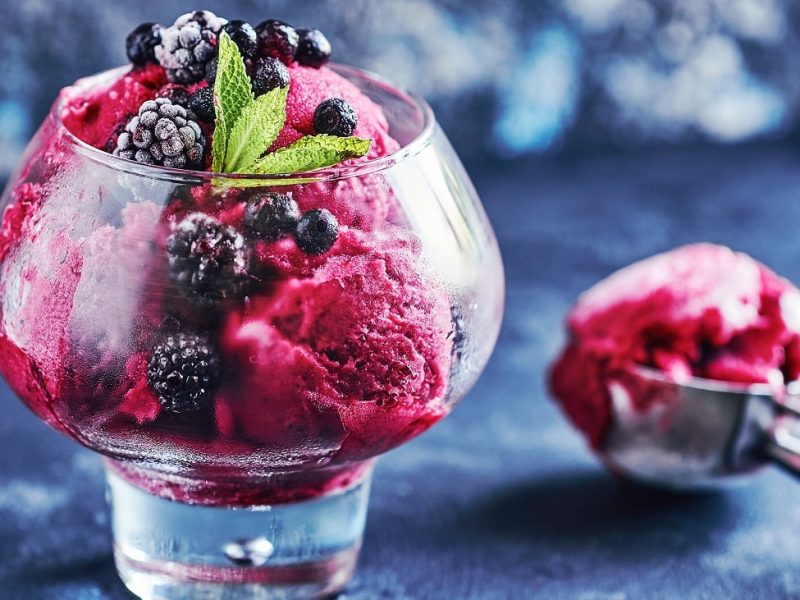 Frontalsicht: Ein Glas mit einem Johannisbeer-Brombeer-Sorbet mit Prosecco. Daneben liegt ein Eisportionierer. der Untergrund ist dunkel.