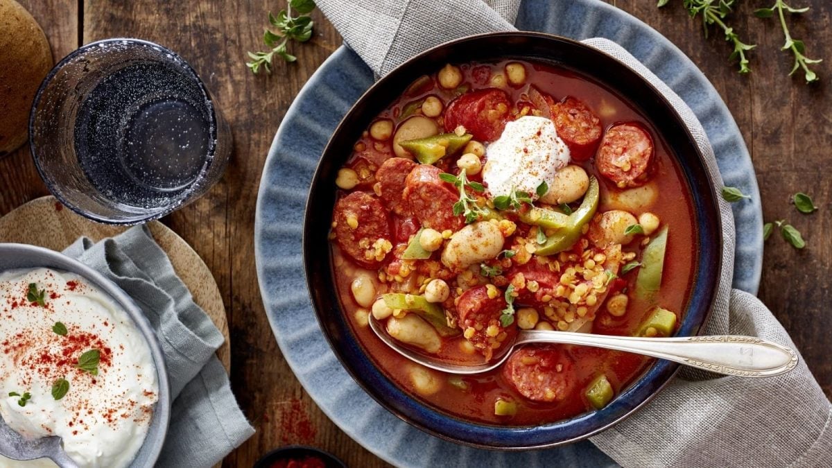 Draufsicht: Blaue Schüssel gefüllt mit einem Eintopf aus Bohnen, Tomaten und Chorizo. Garniert mit saurer Sahne Angerichtet auf einer hölzernen Platte.