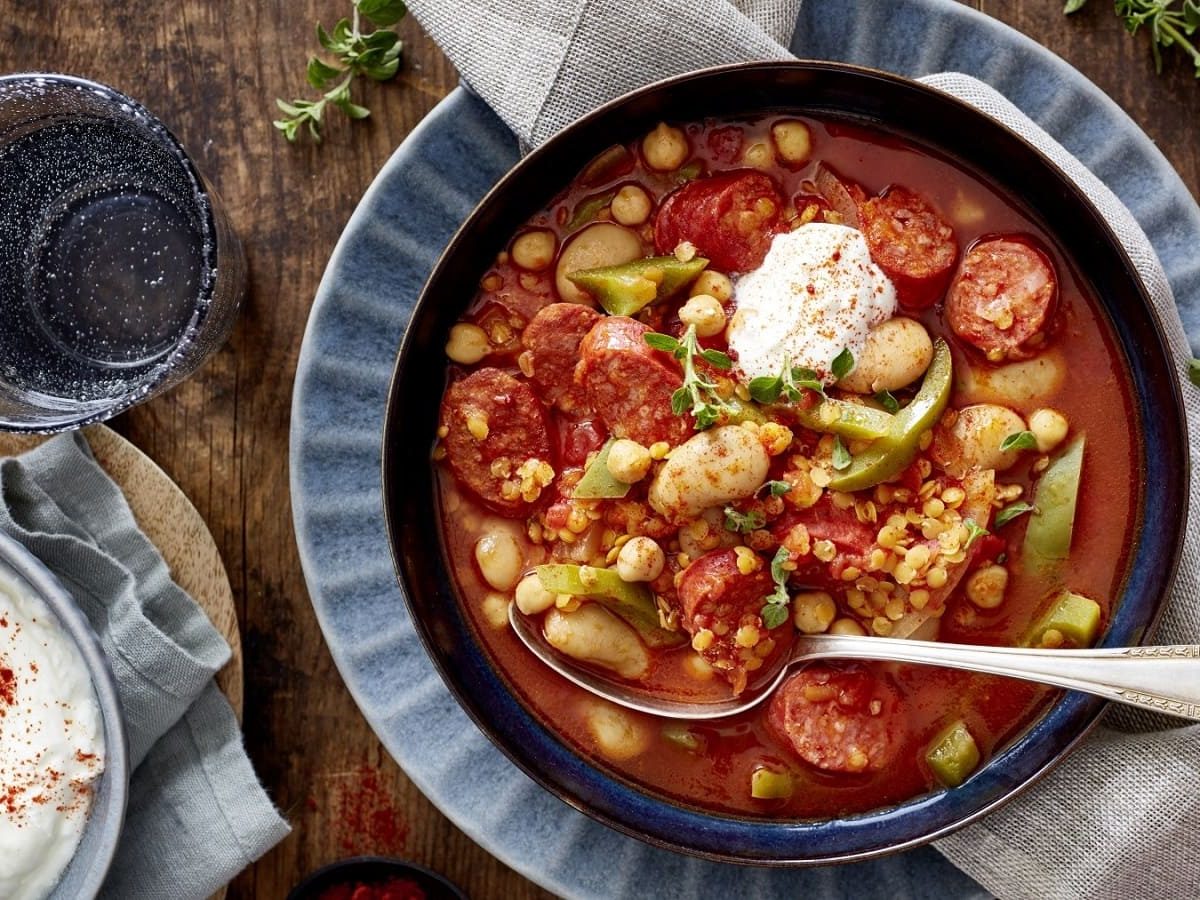 Draufsicht: Blaue Schüssel gefüllt mit einem Eintopf aus Bohnen, Tomaten und Chorizo. Garniert mit saurer Sahne Angerichtet auf einer hölzernen Platte.