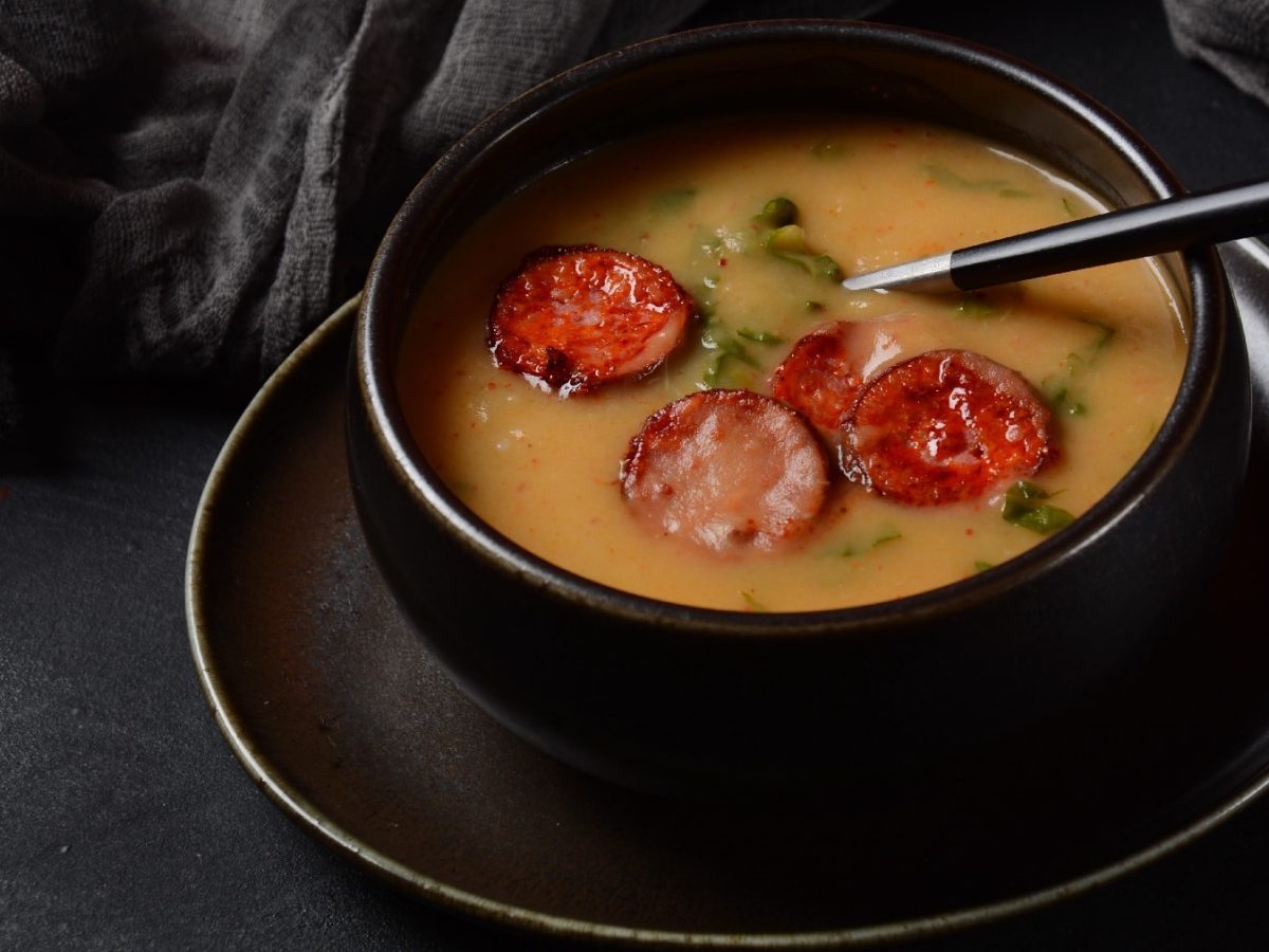 Eine schwarze Schüssel auf dunklem Hintergrund. Darin die Kartoffelsuppe mit ein paar Scheiben gebratener Chorizo.