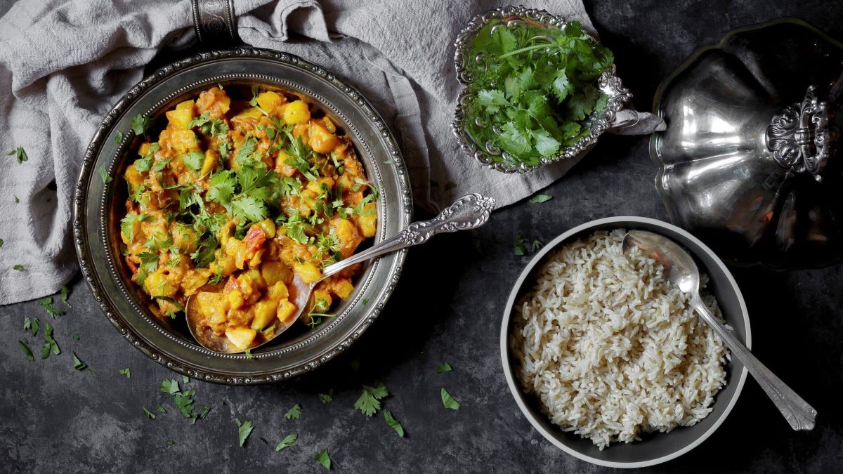 Eine Pfanne Kohlrabi-Curry, daneben eine Schale Reis und eine Schale Koriander. Draufsicht.