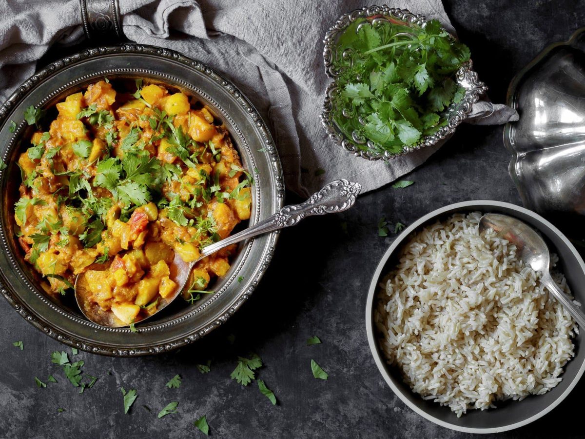 Eine Pfanne Kohlrabi-Curry, daneben eine Schale Reis und eine Schale Koriander. Draufsicht.