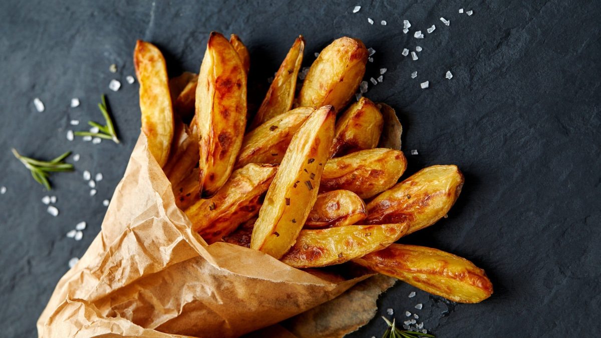 Eine Papiertüte Kohlrabi-Fritten mit Salz und Rosmarin auf dunklem Untergrund.