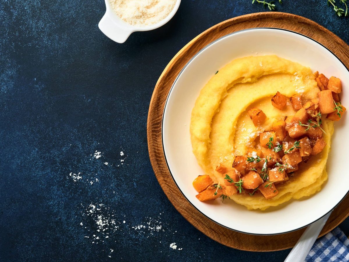 Ein weißer Teller Kürbis-Polenta auf einem dunkelblauem Untergrund. Darüber eine Schale Parmesan und etwas Thymian.