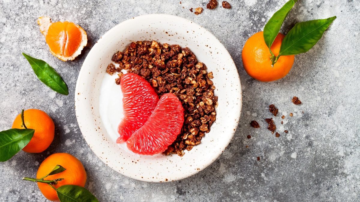 Eine weiße Schale mit Joghurt, pinker Pomelo und dem Lebkuchen Granola auf einem grauen Tisch mit Mandarinen drum herum.