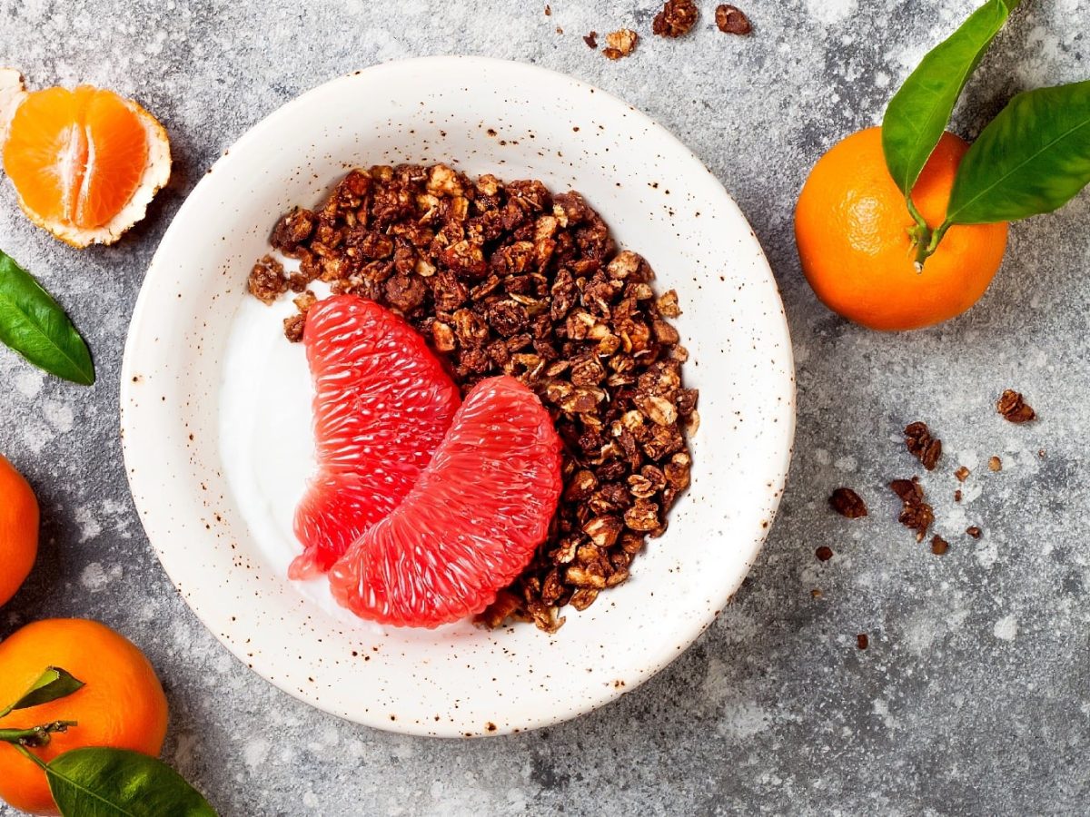 Eine weiße Schale mit Joghurt, pinker Pomelo und dem Lebkuchen Granola auf einem grauen Tisch mit Mandarinen drum herum.